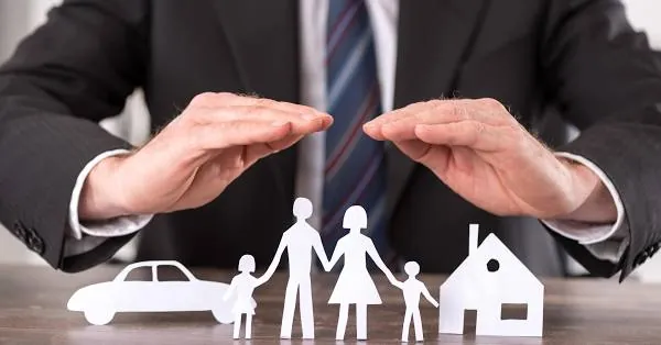 Zwei Hände schützen eine Papierfamilie mit Haus und Auto
